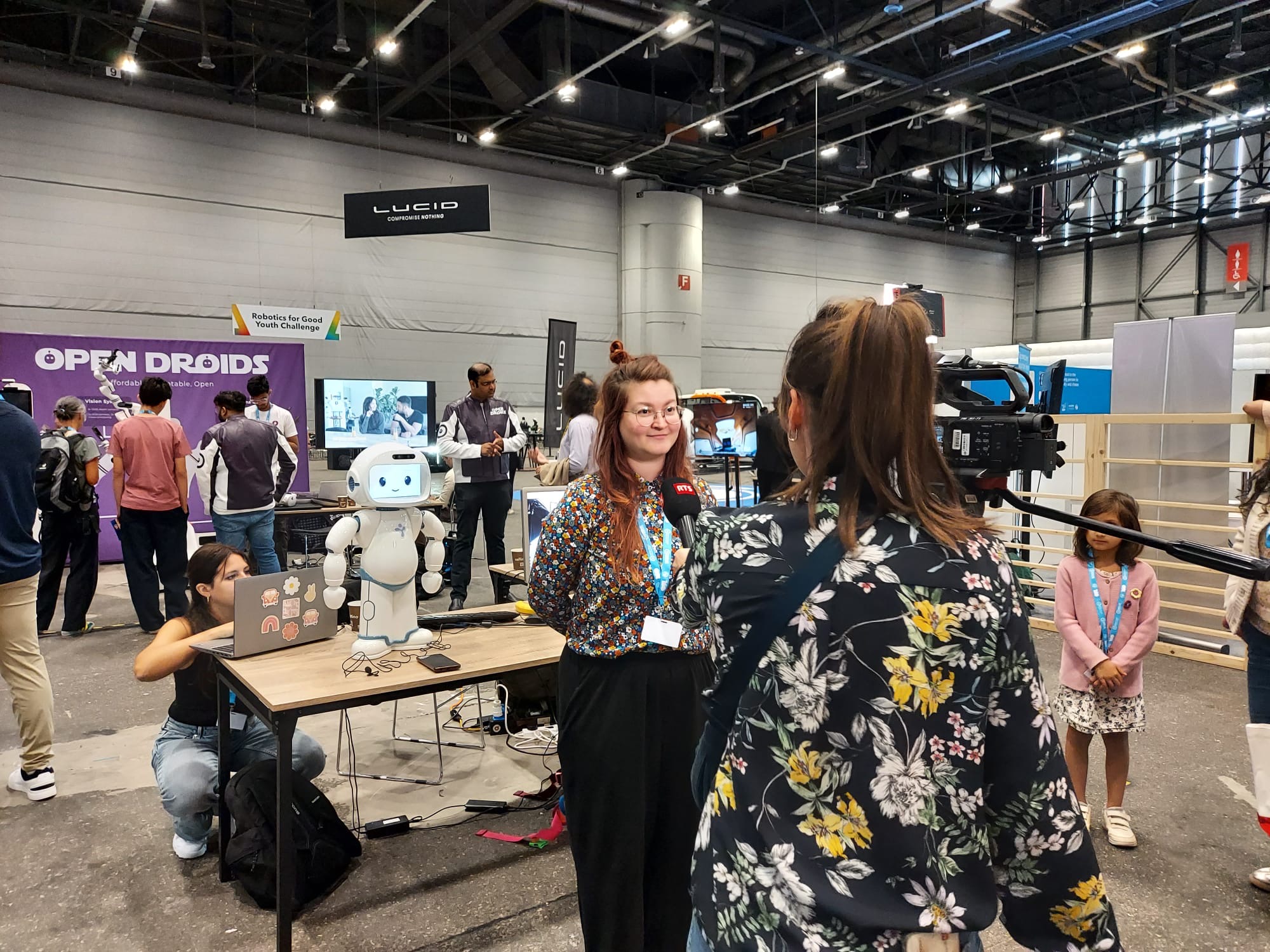 A woman being interviewed for news, with a robot behind her and a microphone being held up to her by a repoter.